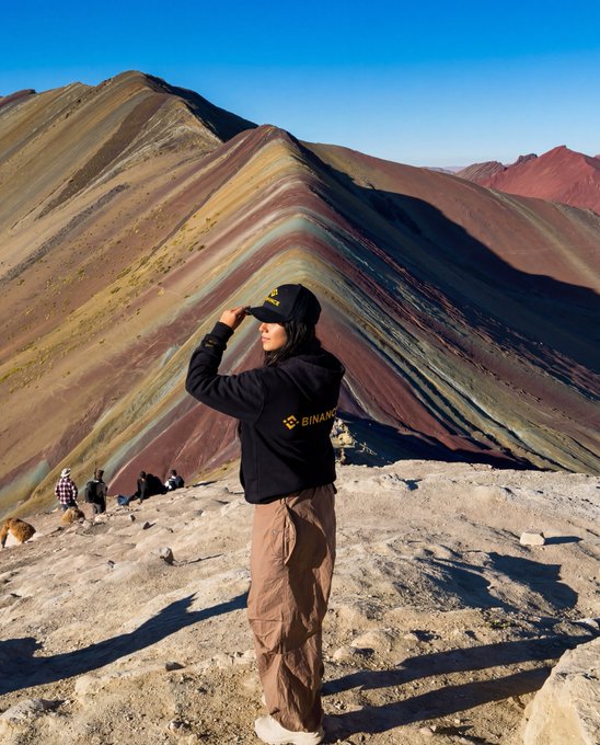A person wearing a black hoodie with the "BINANCE" logo stands atop a colorful, rainbow-striped mountain (Rainbow Mountain/Vinicunca in Peru), enjoying the breathtaking view after a challenging trek. The scene perfectly captures the "Worth the climb" sentiment, showcasing both the natural beauty of the landscape and a subtle nod to the crypto brand worn by the hiker.