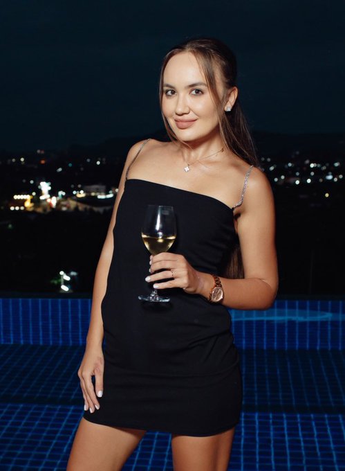 A woman in a black dress holds a glass of white wine while posing by an illuminated rooftop pool at night, with city lights glowing in the background. The image complements the caption's celebratory Friday vibe and playful reference to a "5 figs airdrop," capturing a glamorous, carefree mood as if toasting to a lucky break.