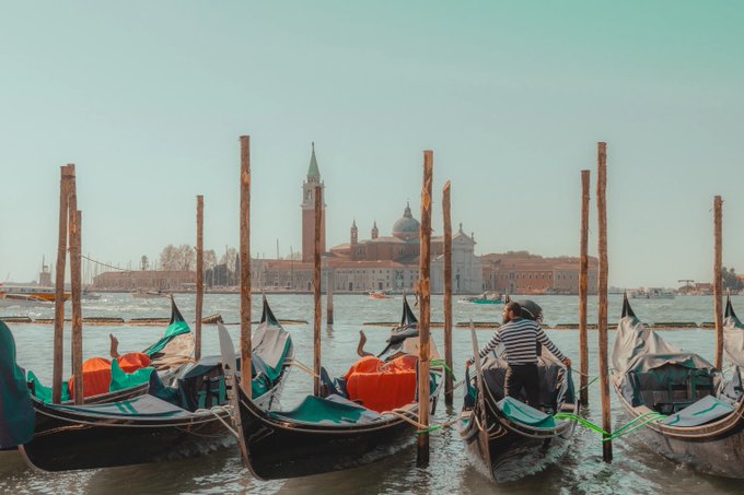 A scenic view of Venice, Italy, showing gondolas docked in the foreground with a gondolier in a striped shirt, and the island of San Giorgio Maggiore with its church and bell tower visible across the water.