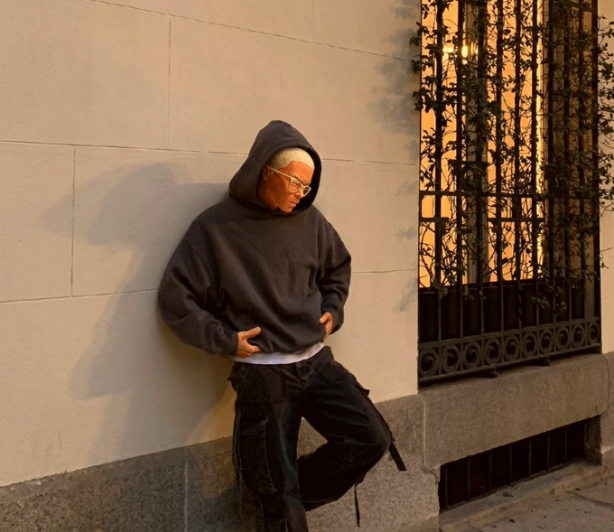 A person wearing a dark hoodie and glasses leans casually against a beige stone wall near a decorative wrought-iron window grate, captured in warm sunset lighting.
