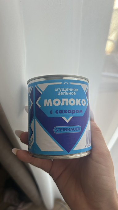 A hand holding a condensed milk can labeled "сгущённое цельное МОЛОКО с сахаром" by the brand Steinhauer, featuring blue and white packaging.