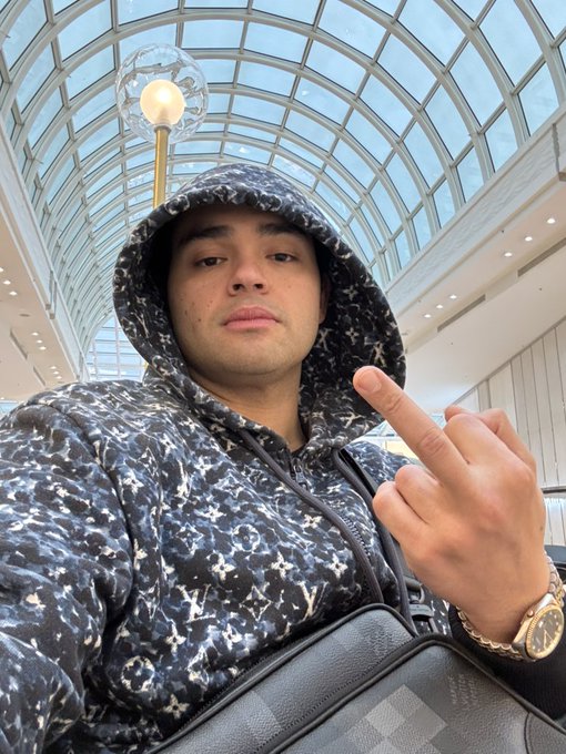 A man in a patterned hooded sweatshirt points his finger at the camera in a building with a glass arched ceiling.