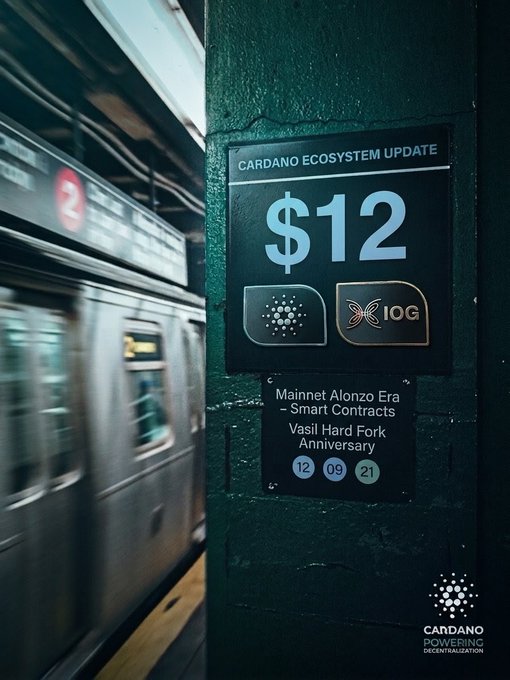 A promotional image for Cardano celebrating a '$12' ecosystem update for the Mainnet Alonzo Era and Vasil Hard Fork Anniversary, set in a subway station with the Cardano and IOG logos.