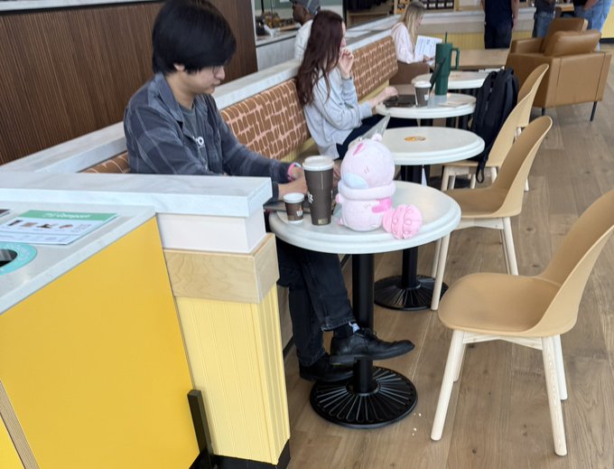 A young man sits alone at a small café table next to a pink plush unicorn, surrounded by empty chairs while using his phone. The scene captures the playful contrast referenced in the post—a major crypto figure casually working amid stuffed toys in a seemingly ordinary setting.