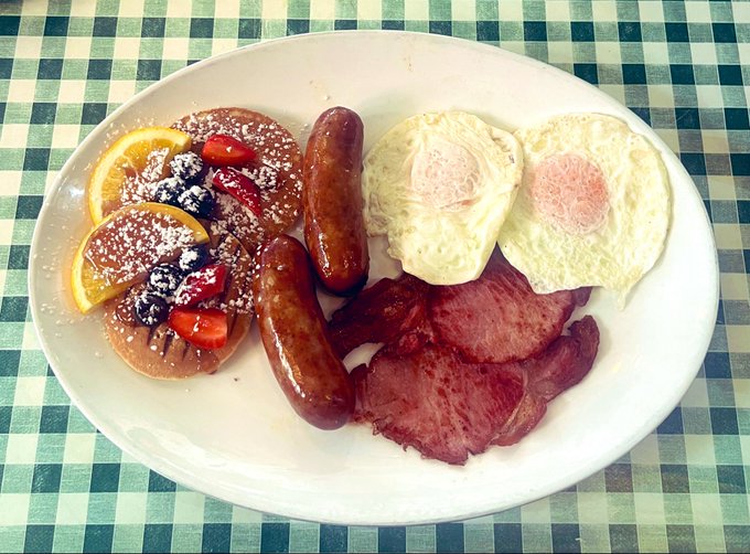 A plate of breakfast food on a green and white checkered tablecloth, featuring pancakes topped with fruit and powdered sugar, sausages, fried eggs, bacon, and orange slices.