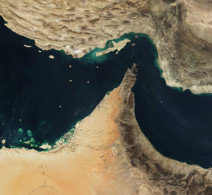 A satellite/aerial view showing the Strait of Hormuz between Iran and the Arabian Peninsula, with the Persian Gulf on the left and the Gulf of Oman on the right, displaying surrounding desert terrain.