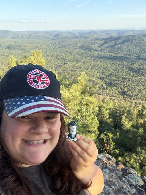 A smiling woman wearing an American flag and Tucson-themed cap holds up a small zombie figurine while posing on a scenic mountain overlook with forested hills in the background.
