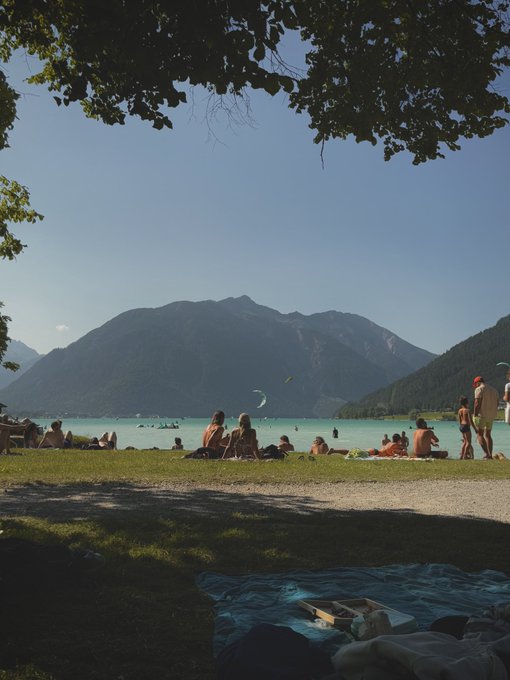 A scenic view of people relaxing on a grassy shore by a lake with mountains in the background.