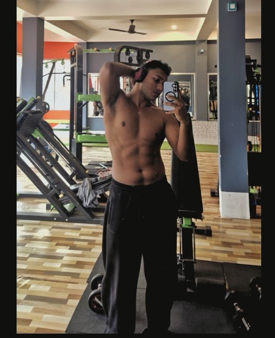 A shirtless, muscular young man takes a mirror selfie at a gym while wearing headphones and black pants.