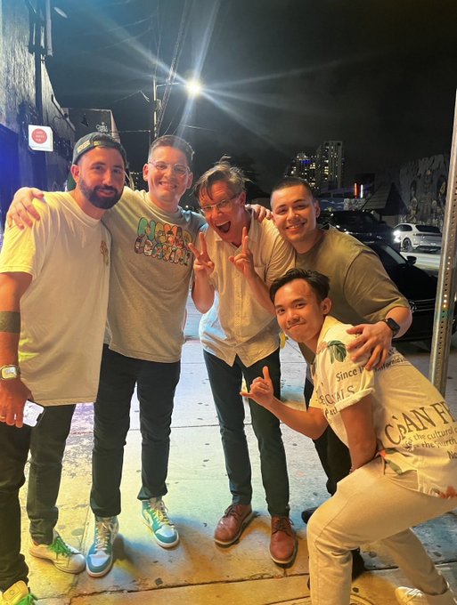 Five men pose together on a city sidewalk at night, with one wearing a 'Nobu' shirt, smiling and making peace signs for the camera.