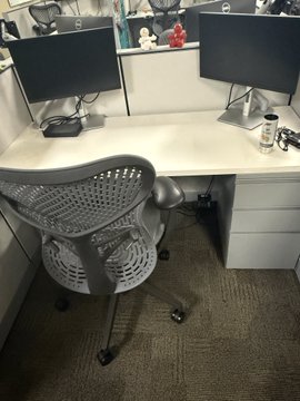 An empty office cubicle workspace features a white desk, dual monitors, and a grey ergonomic mesh chair.