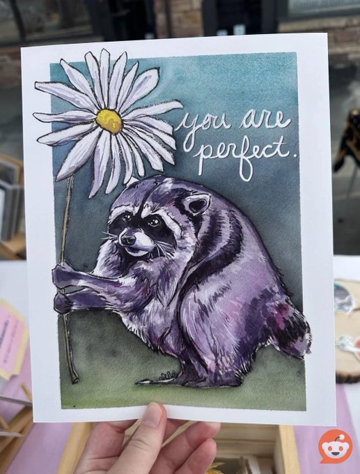 A hand holds up a watercolor art print featuring a detailed illustration of a raccoon hugging a large daisy, with the text "you are perfect." written in cursive next to the flower. The piece beautifully matches the post, showcasing the adorable character "Jimothy" the raccoon receiving a heartfelt compliment.