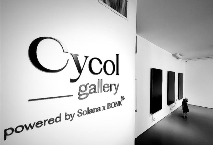 A black-and-white photo of the interior of Cycol Gallery, showing its wall logo reading "Cycol gallery, powered by Solana x BONK," with a person viewing the artwork on display — illustrating the gallery's brick-and-mortar renewal under its Solana and Bonk partnership.