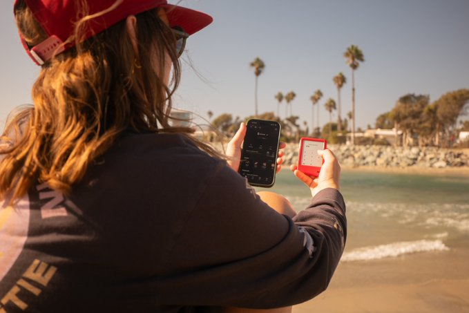 A person wearing a red cap and dark hoodie sits at a sunny beach, holding a smartphone in one hand and a small red compact device in the other, with palm trees lining the shore in the background.