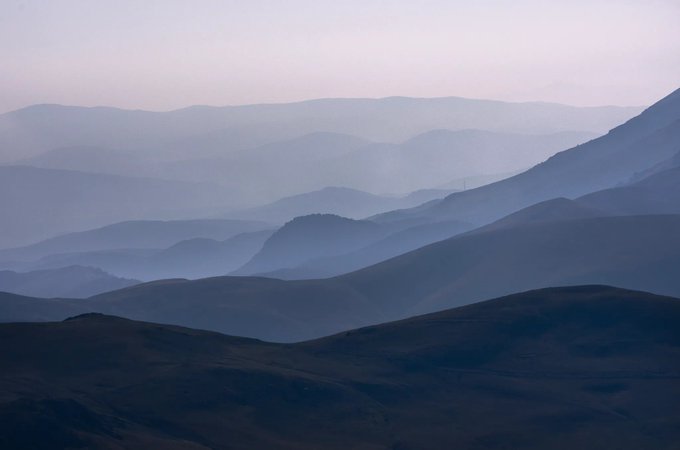 A serene landscape photograph showing layered silhouettes of rolling hills and mountains fading into a hazy, muted purple-blue atmosphere.