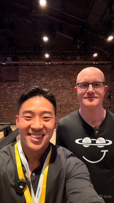 The image shows two men standing next to each other, both wearing black shirts with white designs. One of them is holding a camera, possibly taking a selfie or recording something interesting happening around them. The background features a brick wall and a stage setup, suggesting that they might be at an event or gathering. There are also some text elements in the image, including "Grok has money SpaceX owns that money $DRB". This could indicate that the men are attending an event related to space exploration or technology, possibly involving companies like SpaceX.