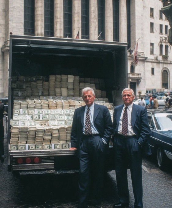 Two men in suits standing in front of a truck loaded with stacks of cash on a city street, with classical architecture and an American flag in the background.