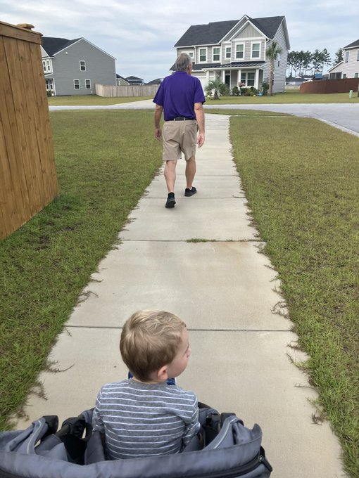 A young child sits in a stroller on a sidewalk, watching an older man in a purple shirt and khaki shorts walk ahead of them toward a residential neighborhood.