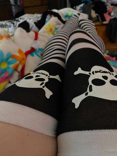 A close-up view of legs wearing black and white striped thigh-high socks featuring a large white skull and crossbones design, resting on a colorful floral blanket.