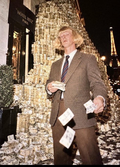 A blonde man in a brown houndstooth suit stands in front of the Hotel Ritz Paris with a massive pile of money stacked behind him, holding cash in his hands as bills fall through the air, with the Eiffel Tower visible in the background.