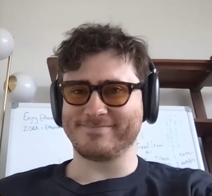 A man with curly brown hair, glasses, and headphones smiles at the camera, with a whiteboard behind him containing notes that include 'ZORA' and 'Feed Token'.