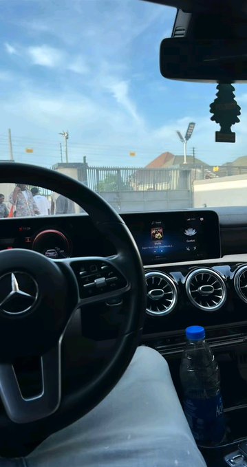 Interior view from the driver's seat of a Mercedes-Benz, showing the steering wheel with the logo, dashboard controls, and a water bottle in the cup holder.