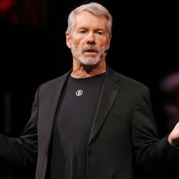 A middle-aged man with grey hair and a beard, wearing a black blazer over a black t-shirt with a circular pendant, is captured mid-speech on a dark stage.