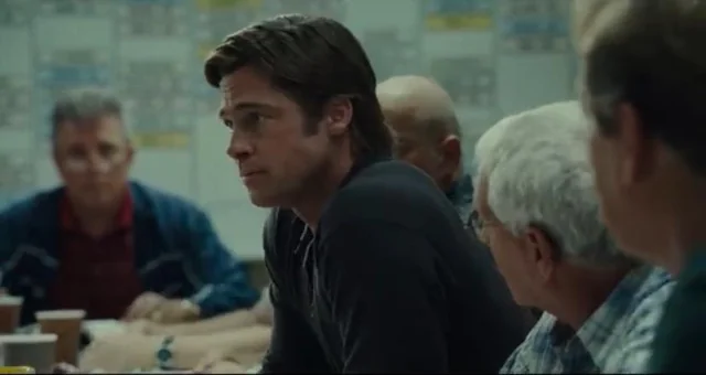 A scene from a movie featuring a young man with dark hair in a suit sitting at a table with several older men in what appears to be a diner or office setting.