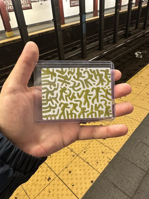 A hand holds a small clear plastic case displaying a card with a yellow and white squiggle pattern, photographed on a subway platform with yellow tactile warning strips and tracks visible in the background.