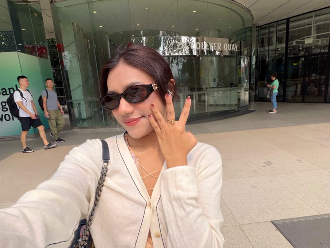 A woman wearing sunglasses and a white cardigan poses for a selfie in front of the Tower Quays building, holding up her fingers in a gesture.