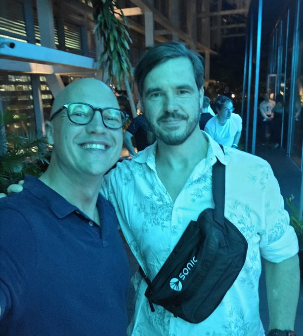 Two men pose for a photo at night; the man on the right has a beard and wears a white patterned shirt with a black 'sonic' fanny pack.