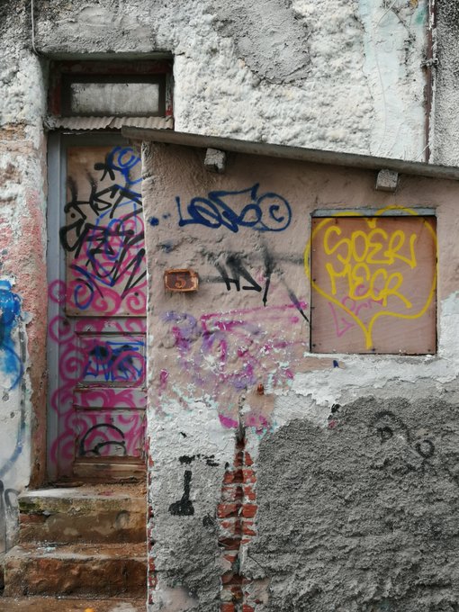 A weathered urban doorway and adjacent boarded-up window covered in colorful graffiti tags, including a yellow 'GOZI BBR' tag and a blue 'LOKO' tag on the pink-tagged door.
