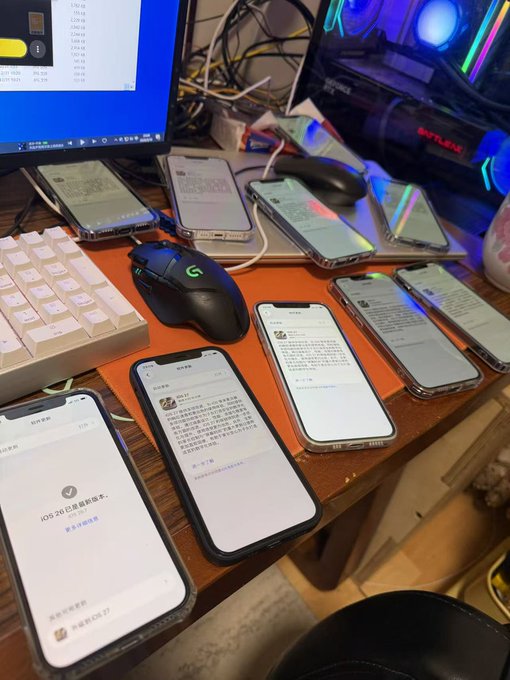 A wooden desk with multiple iPhones laid out, all appearing to be in a state of testing or setup, with a monitor displaying file lists, a Logitech mouse, and a keyboard.