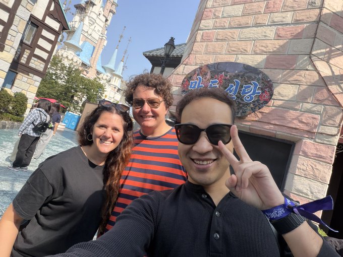 Three people pose for a selfie at what appears to be a Disney park, with one making a peace sign near a Korean sign that reads '마인 신발' (Mine Shoes).