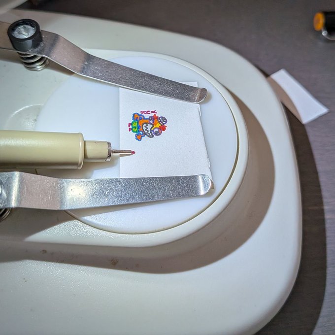 A close-up of a machine with metal arms using a small pen-like tool on a square white paper, appearing to draw or write a colorful small design.