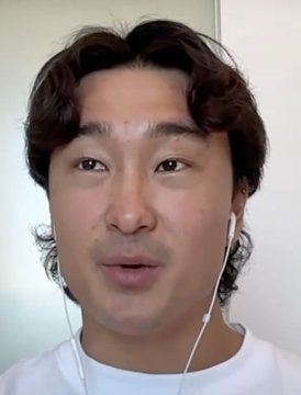 A close-up selfie of a young man with wavy dark hair wearing a white shirt and white earphones against a plain wall.