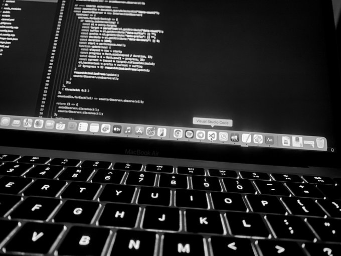 A close-up black and white photo of a MacBook Air displaying code in Visual Studio Code, with the macOS dock visible at the bottom of the screen.