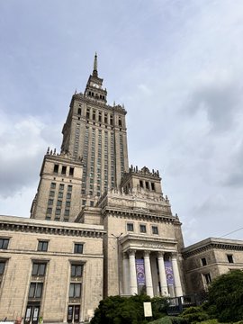 The image features a tall building with multiple levels, possibly an old stone structure or a modern skyscraper. The sky above appears cloudy, suggesting that the photo was taken during the day. There are several people visible in the scene, some of whom might be attending the Bitcoin Film Festival held at this location. A car can also be seen parked nearby, likely belonging to one of the attendees.