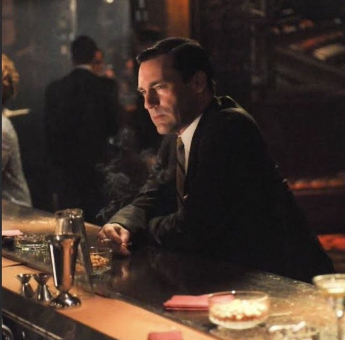 A man in a dark suit sitting alone at a dimly lit bar, staring thoughtfully with a cigarette in hand.