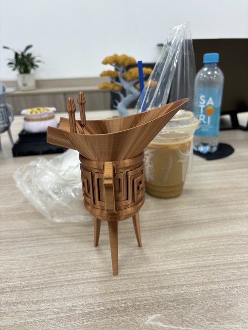 A wooden decorative figurine shaped like a traditional tripod vessel (similar to a Chinese ding or jue) sits on a wooden table alongside a clear plastic cup of iced coffee and a bottle of water.