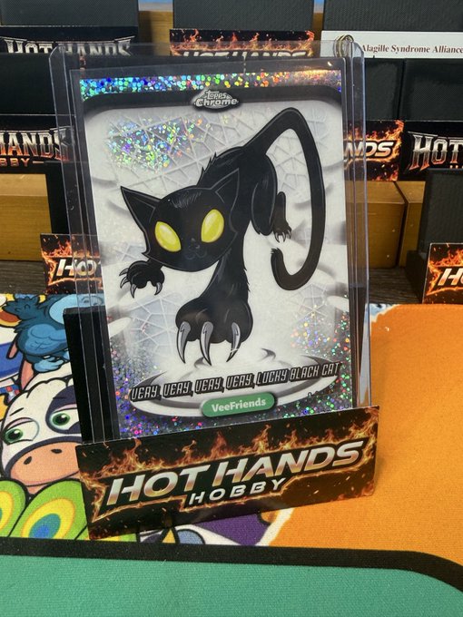 A black cat-themed trading card labeled "Lucky Black Cat" from the "VeeFriends" series is displayed on a stand with "HOT HANDS HOBBY" branding, featuring a holographic shimmer background.