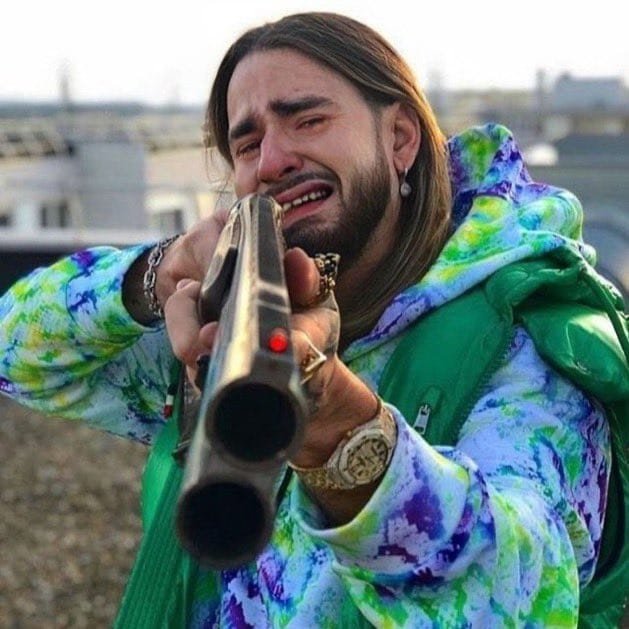 The image shows a tearful man with long hair, wearing a colorful tie-dye jacket and gold watch, crying while aiming a large gun at the camera. The caption should reference the post's context: "@waleswoosh anytime Kaito makes an announcement without paying Evelyn rewards"

Suggested caption: "Crying dramatically with a gun aimed at the camera, this is the exact energy of getting hyped for a Kaito announcement only to realize the Evelyn rewards never came through 💔🔫"

Alternative shorter version: "When you're emotionally devastated waiting for Evelyn rewards that never come after a Kaito announcement 😭🔫"