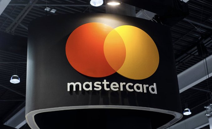 The image features a large Mastercard logo hanging from the ceiling at an indoor event or exhibition, with the text "MASTERCARD RECEIVES NEW YORK BITLENSE" above it. The company has recently received approval for its New York BitLicense, which is a major step in their push towards digital asset payment infrastructure. This license allows Mastercard to operate more securely and efficiently within the financial services industry.