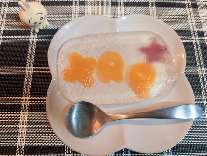 A piece of jelly cake spelling "XQQ" with a star shape, served on a pink bear-shaped plate with a spoon, next to a small bunny figurine on a checkered mat.
