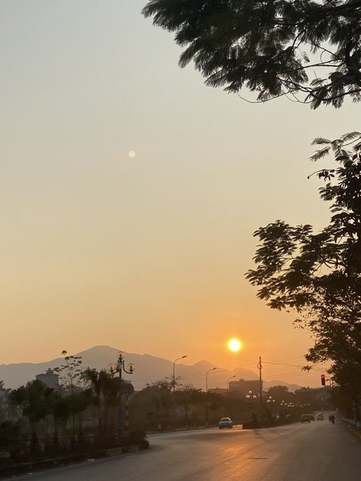 A scenic sunset view over a road with mountains in the distance, silhouetted trees framing the sky, and a few cars driving along the street.