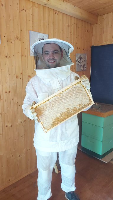 Photo of a smiling man wearing a full white beekeeping suit and protective veil, holding up a wooden frame covered in honeycomb inside a wooden cabin with a green beehive box in the background.
