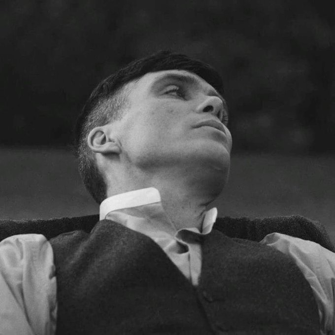 A black and white cinematic portrait of a young man wearing a vest over a white shirt with an upturned collar, gazing upward into the light with a contemplative expression.