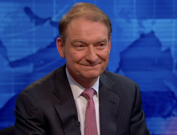 SEC Chair Paul Atkins smiles during an interview, appearing in response to news about his statement that the US must lead in AI, crypto, and financial innovation by advancing the CLARITY Act. He is dressed in a dark suit with a red patterned tie, set against a blue world map backdrop typical of a news studio.