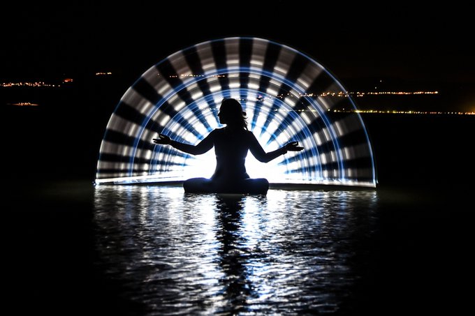 A silhouette of a person meditating in lotus pose on water at night, illuminated from behind by a glowing, striped semicircular light display, with city lights visible on the distant horizon.