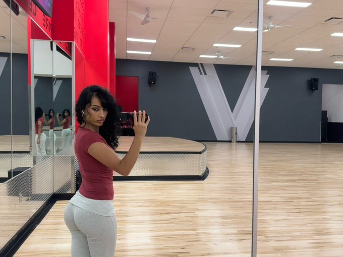 A woman in a maroon top and gray leggings takes a mirror selfie with her phone in a dance studio featuring wooden floors and a stage, with a large "V" graphic visible on the back wall.